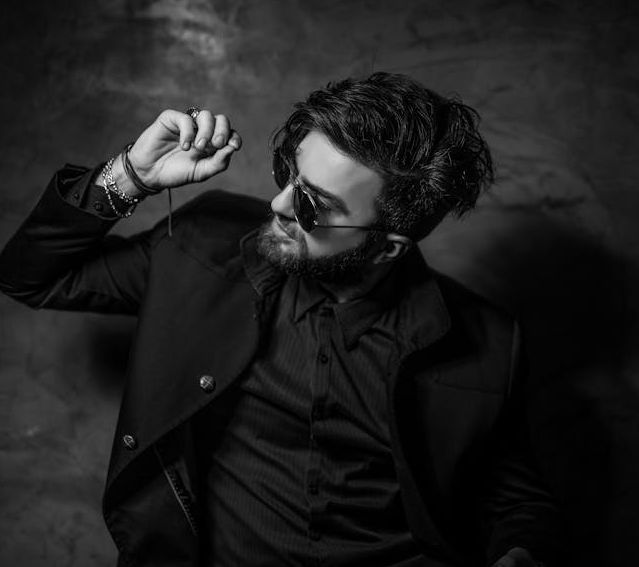 Black and white portrait of a fashionable man in sunglasses and suit posing confidently indoors.